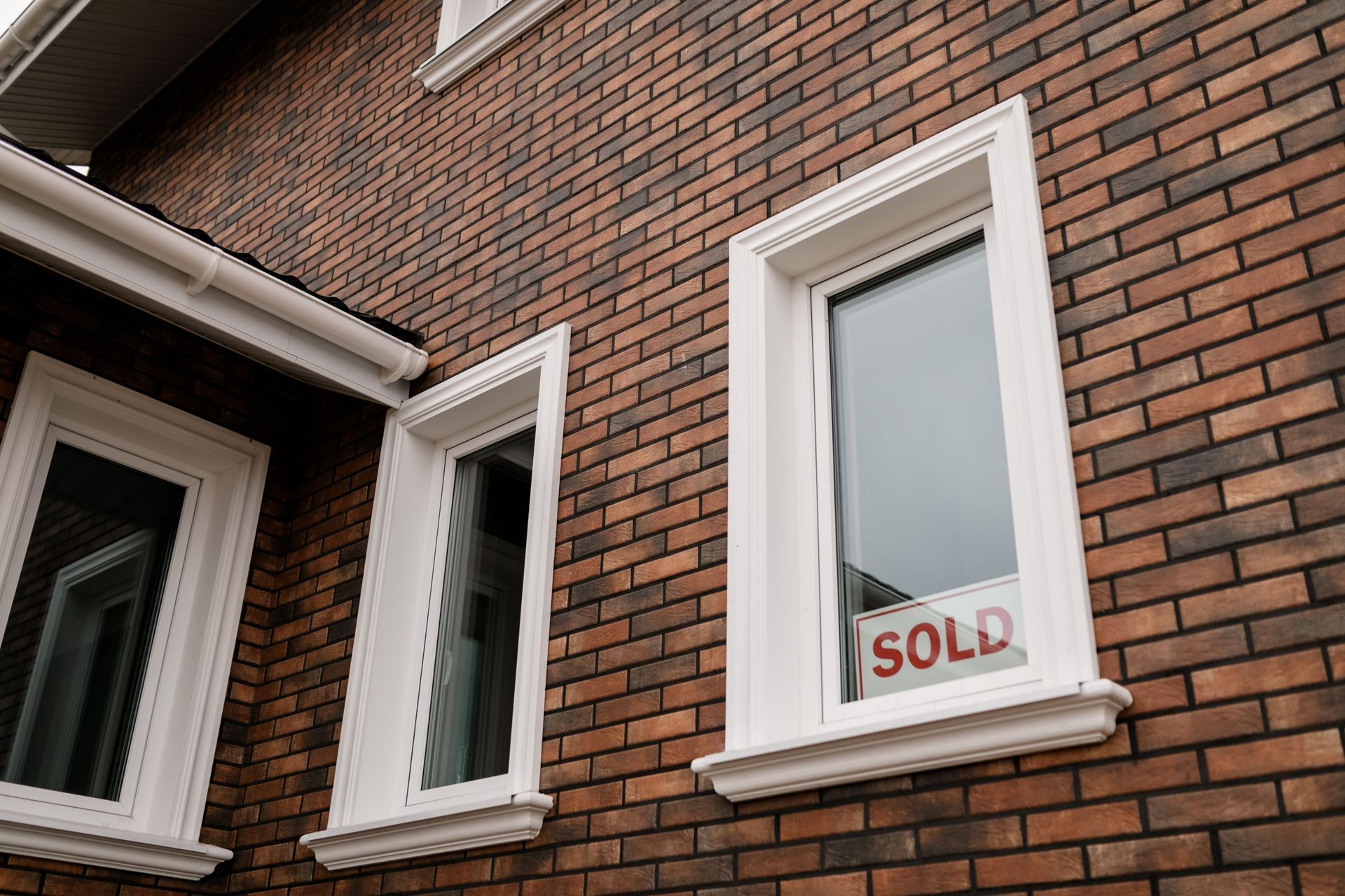 Brick house exterior with white-trimmed windows and a red “SOLD” sign in one window