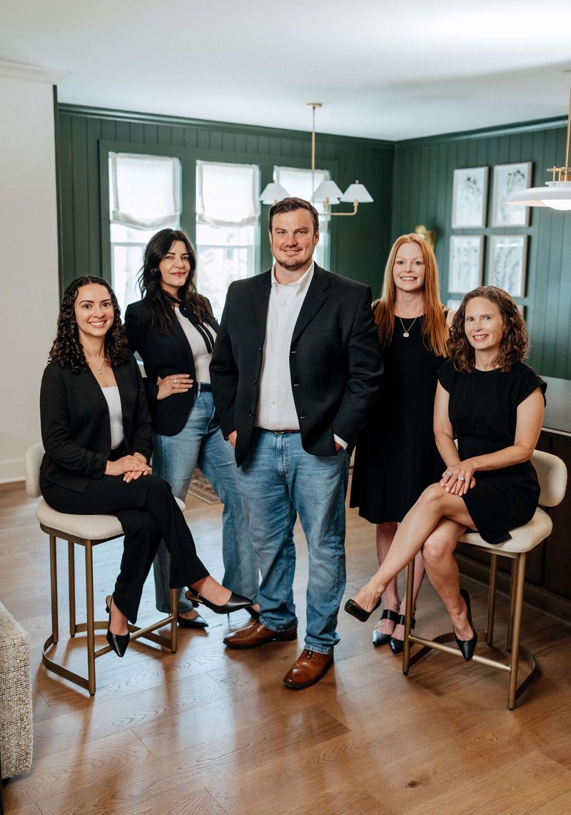 Five adults posing in a living room, with gray walls, large windows, and wood floors.