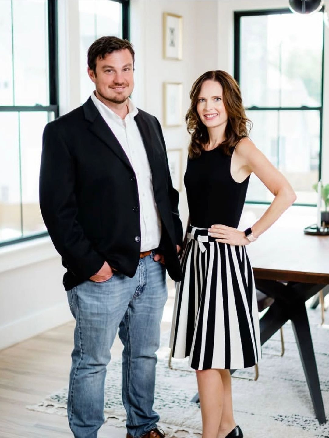 Two people standing in a bright room, one in a black suit and one in a black top and striped skirt.