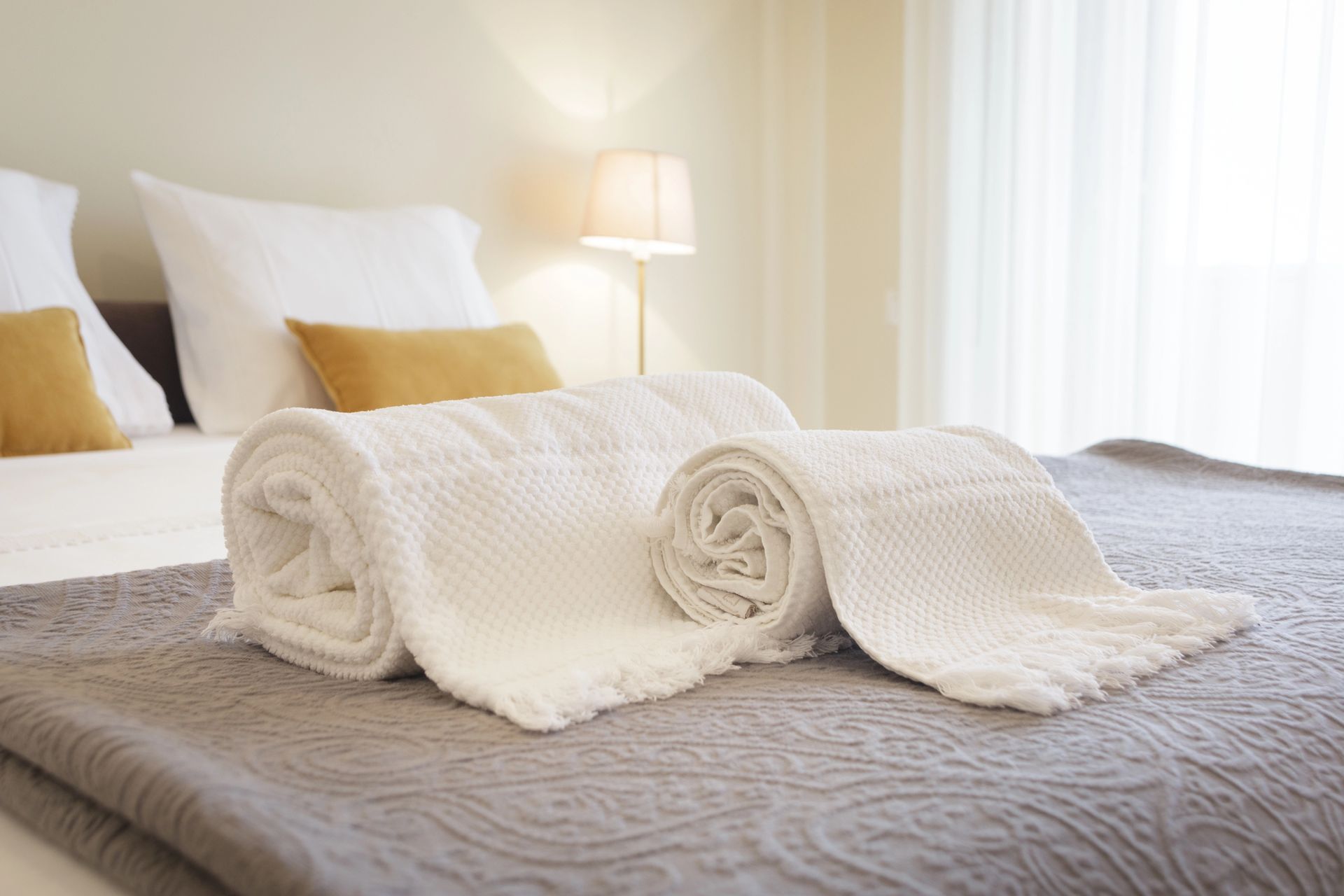 Asciugamani bianchi arrotolati su un letto ben rifatto con una coperta grigia e una lampada da comodino dalla luce soffusa.