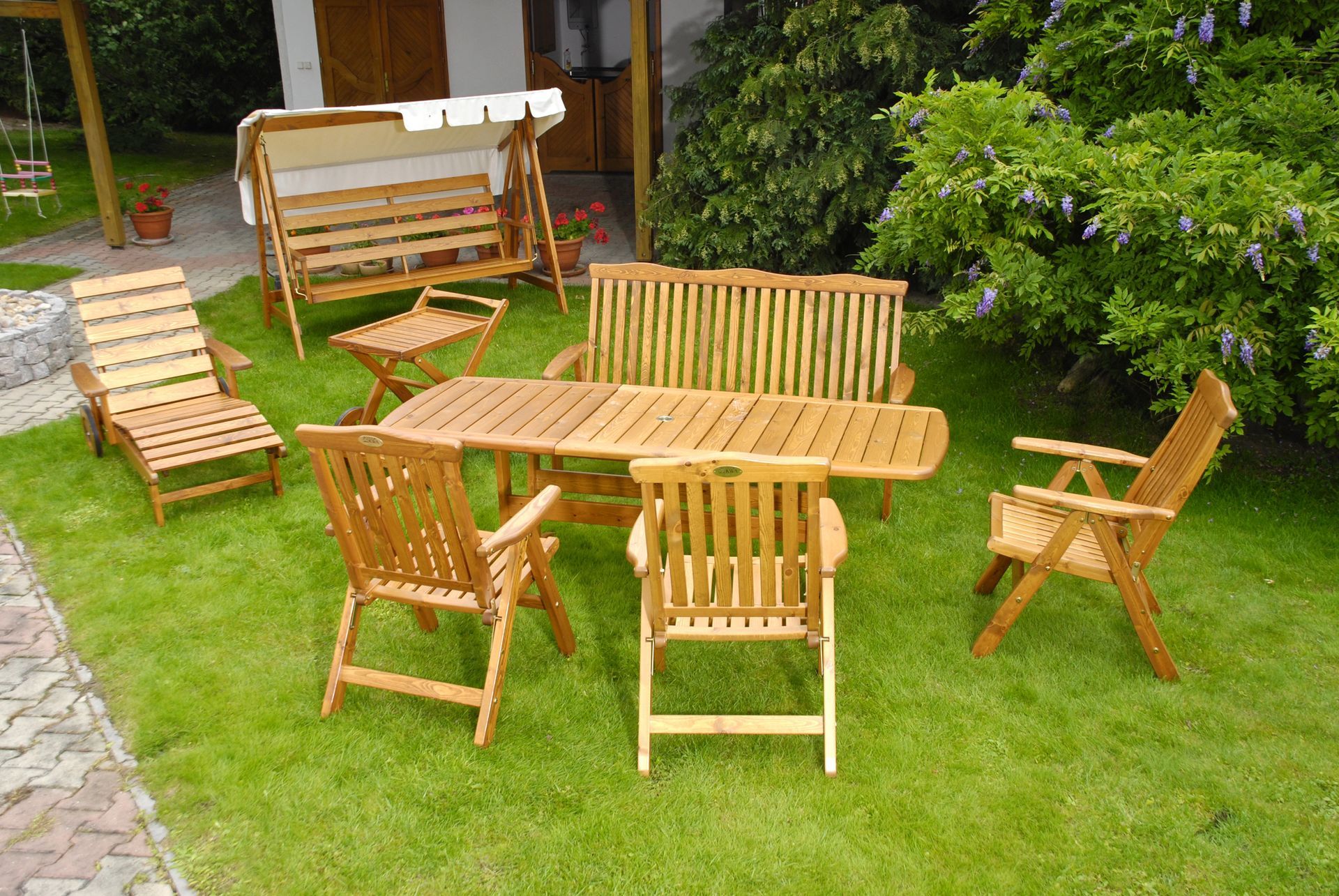 Tavolo e sedie da patio in legno su un prato erboso accanto a una panchina da giardino e vegetazione.