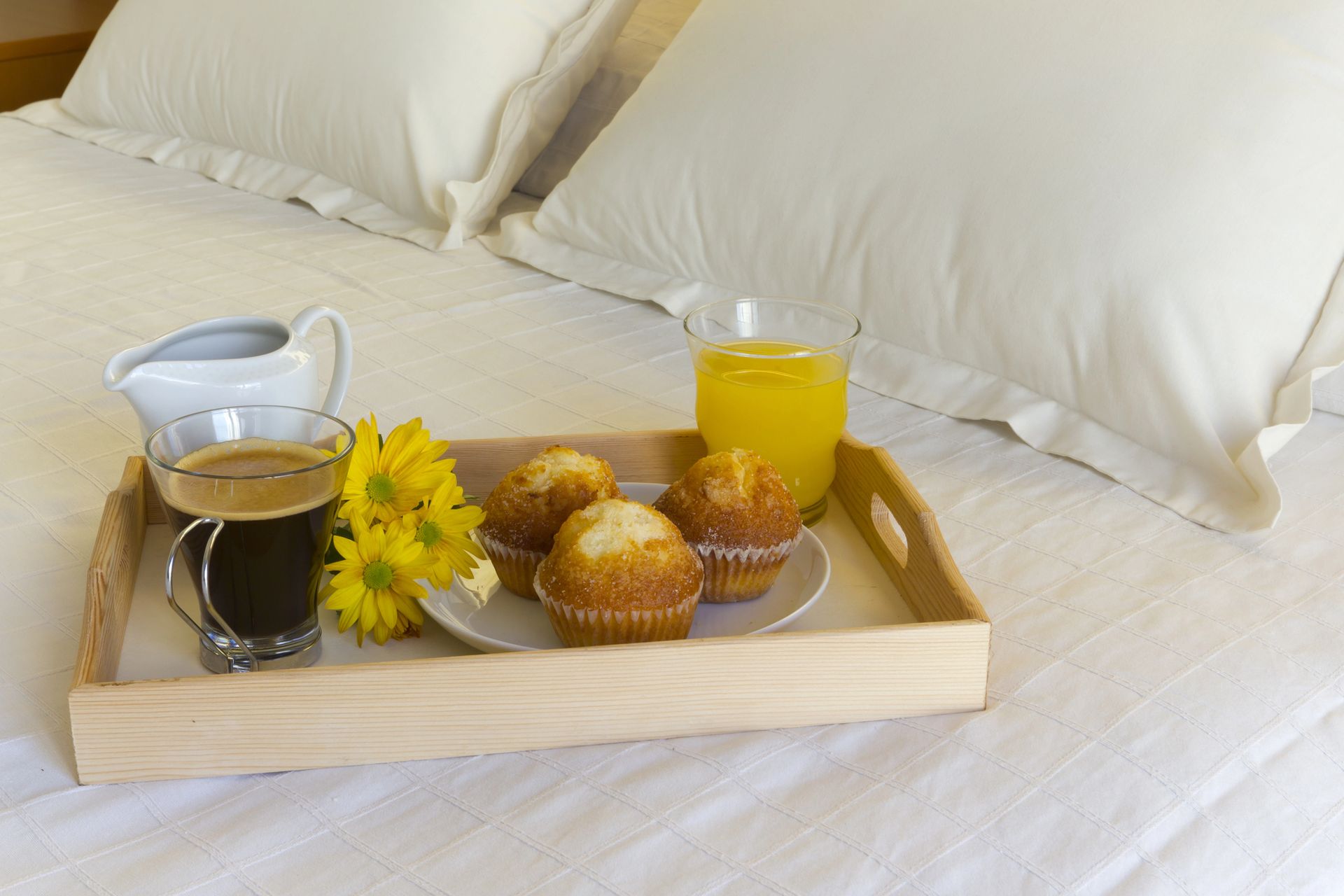 Vassoio per la colazione con caffè, succo di frutta, pasticcini e fiori gialli su un letto bianco.