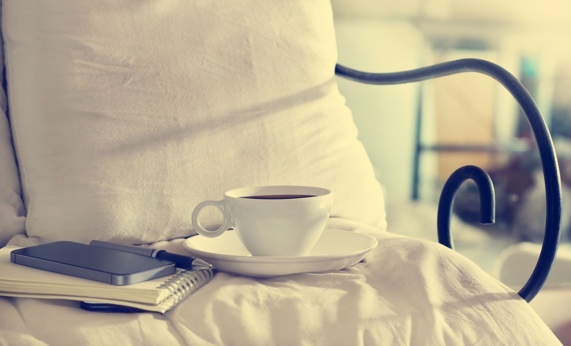 Tazza e piattino bianchi su un letto accanto a un quaderno chiuso e una penna su un cuscino color crema in una luce soffusa.