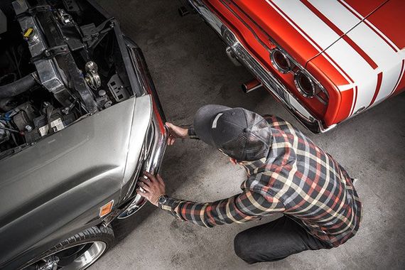 Mechanic kneeling between two cars, checking the front bumper of a silver car in a garage.
