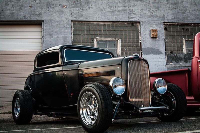 Black vintage hot rod parked beside a red truck against a gray brick wall.