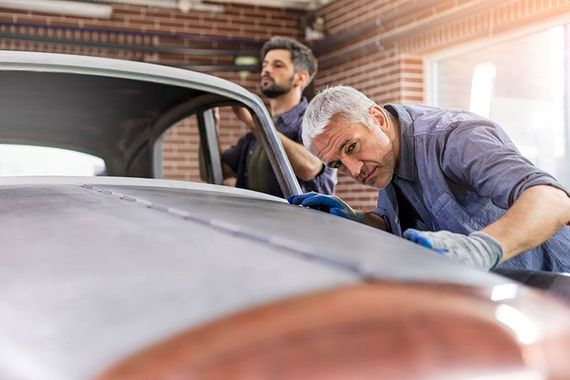 Two mechanics inspecting a car hood in a garage, one leaning over the engine.