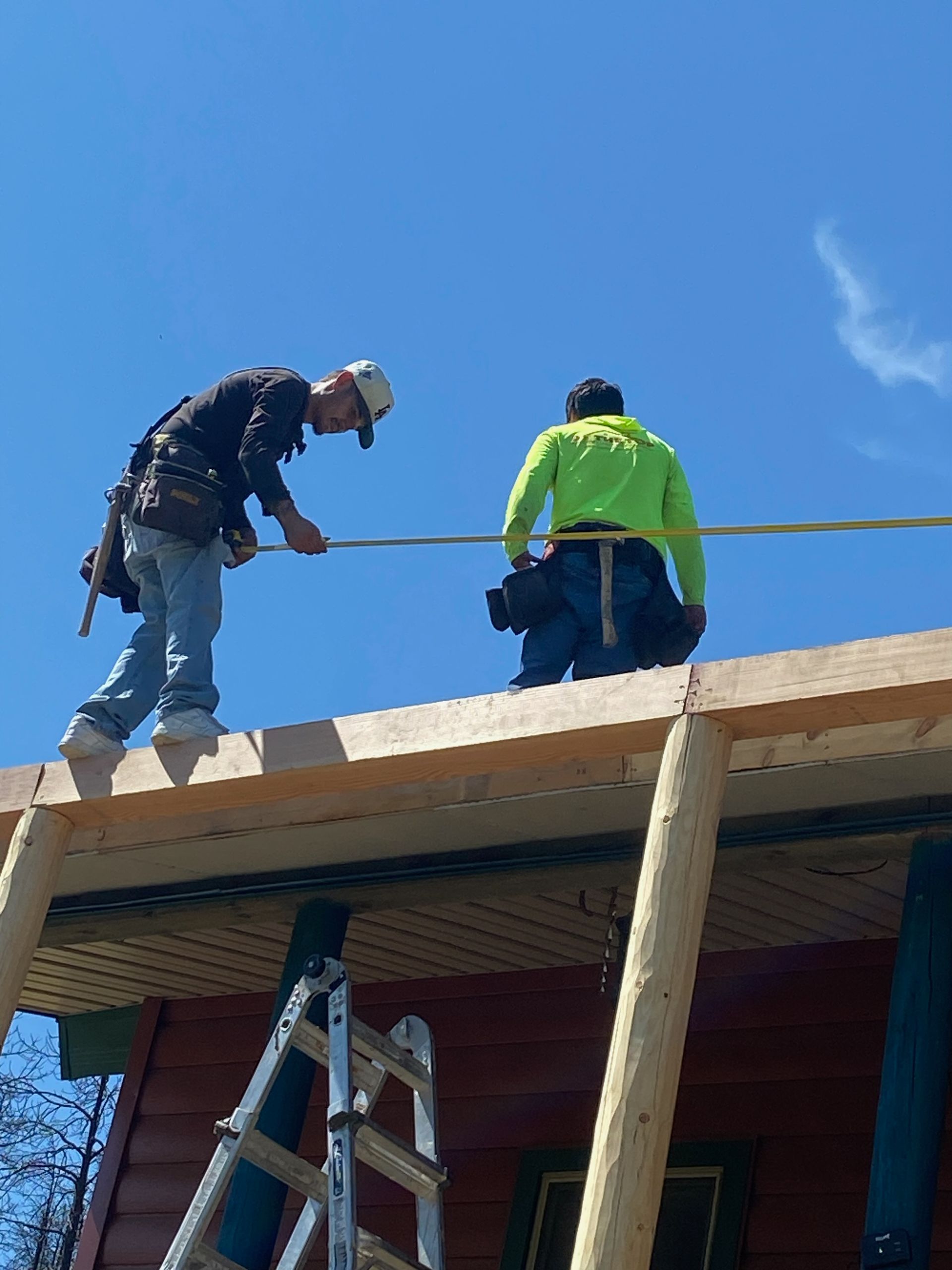 Two construction workers on a roof, one in jeans, one in neon green, working in sunlight.
