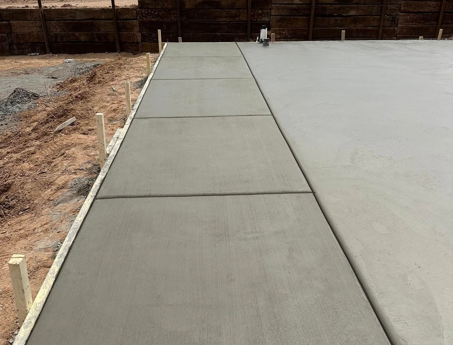 Freshly poured concrete walkway with expansion joints, next to dirt and a fence.