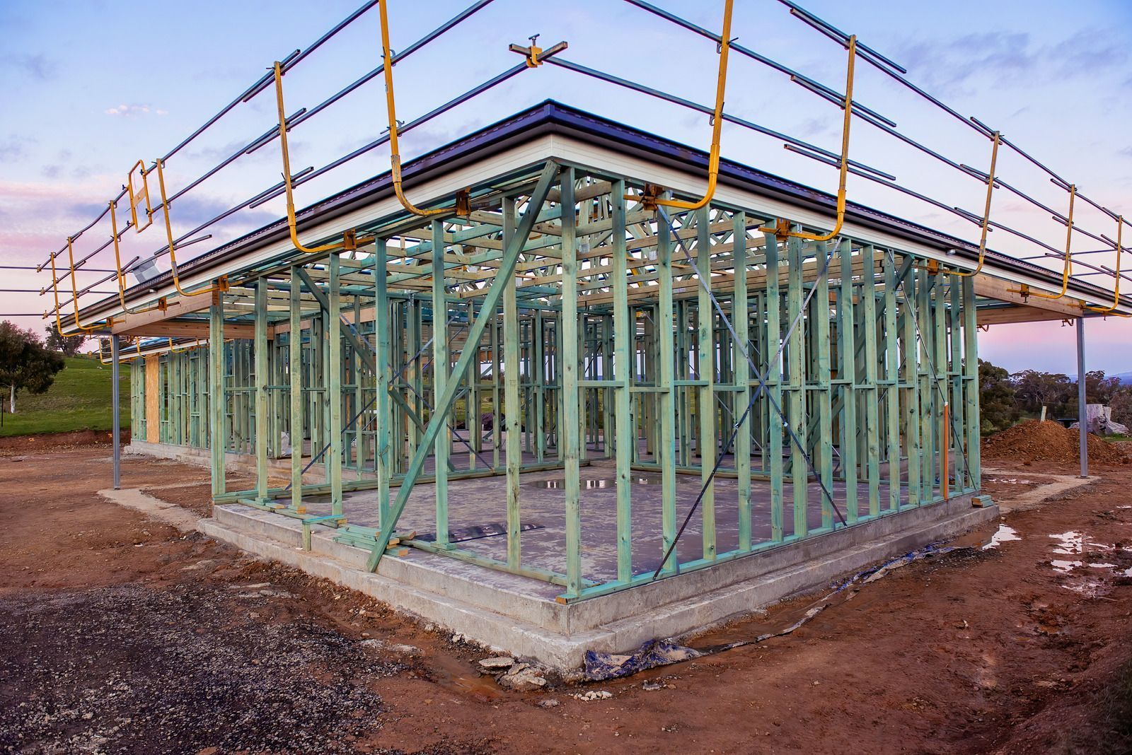 Wooden frame of a house under construction with scaffolding, set on a concrete foundation, outdoors with a blue sky.