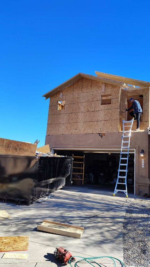 Construction of a two-story building with a garage. A worker on a ladder adds siding. Blue sky, a dumpster, and tools.