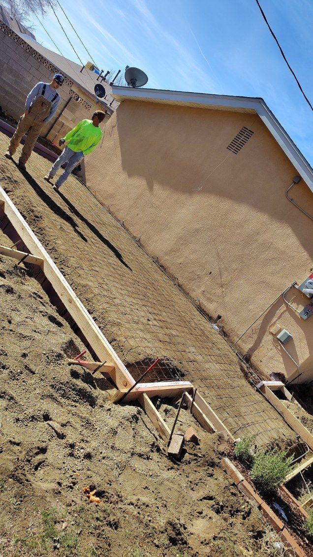 Two men by a house on a hillside, supporting a wooden structure. Exterior shot, sunny day.