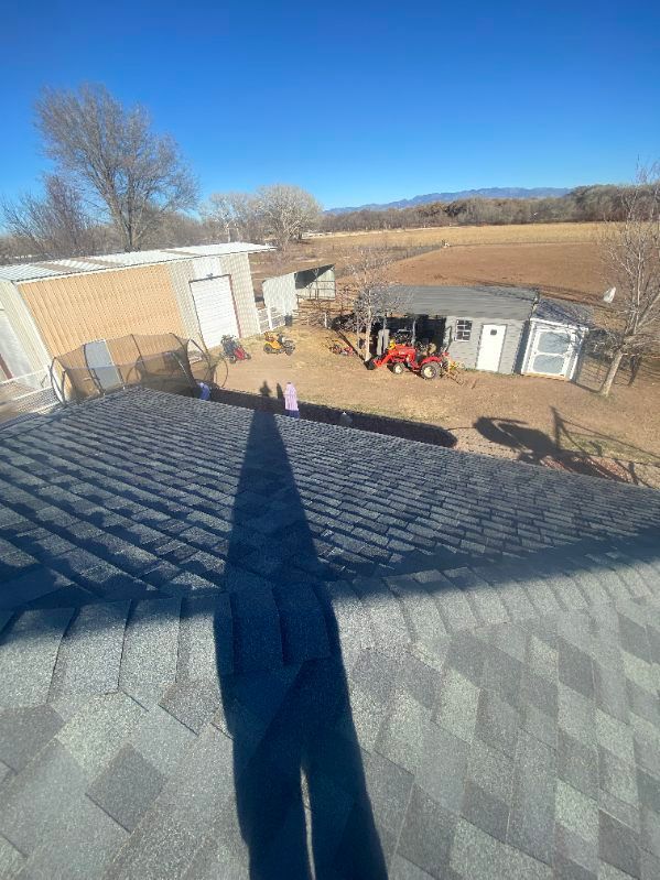 View from a roof of a house with dark gray shingles overlooking a yard with buildings and a tractor.