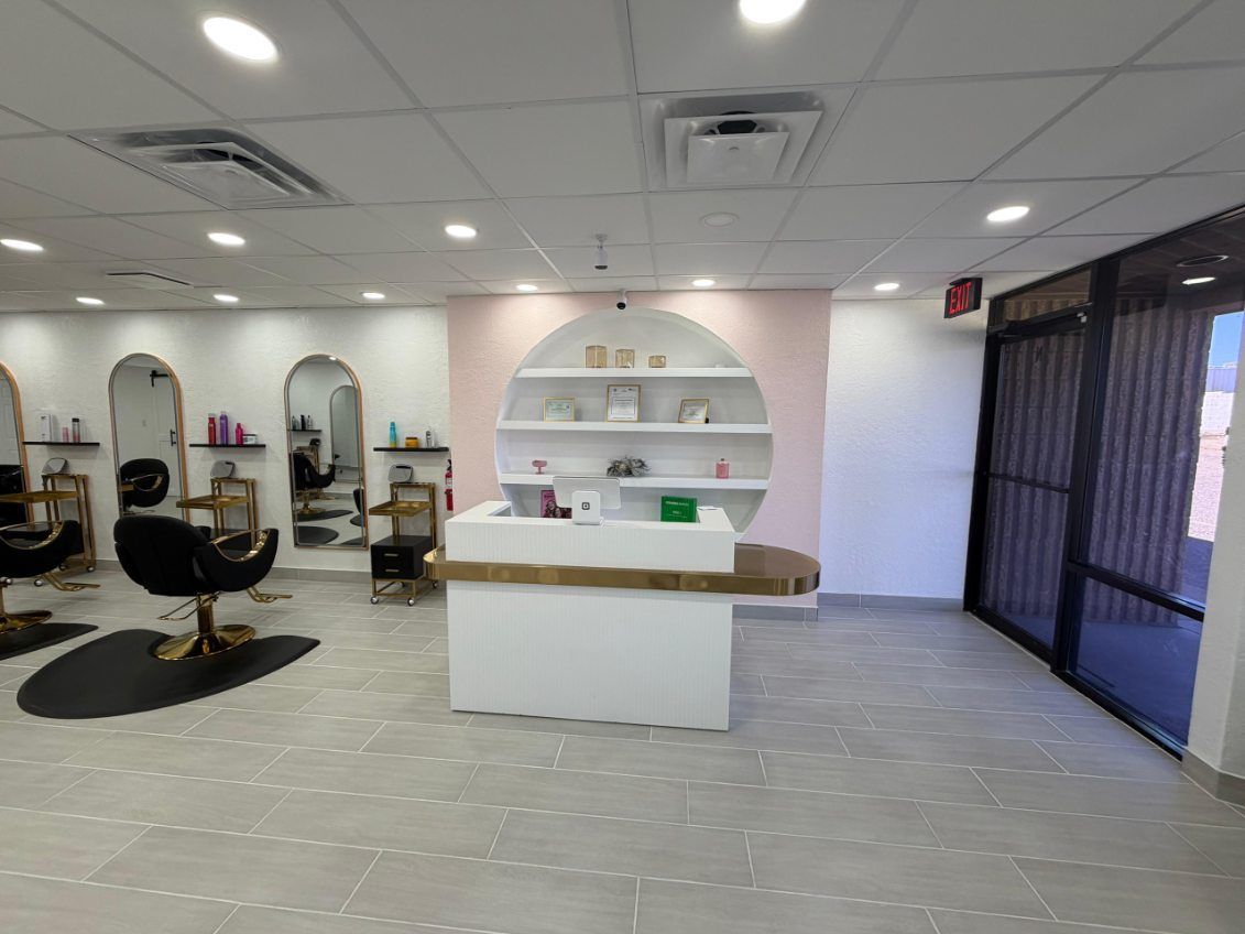 Interior of a hair salon with white walls, mirrors, chairs, and a front desk. Light pink accent wall with shelving.