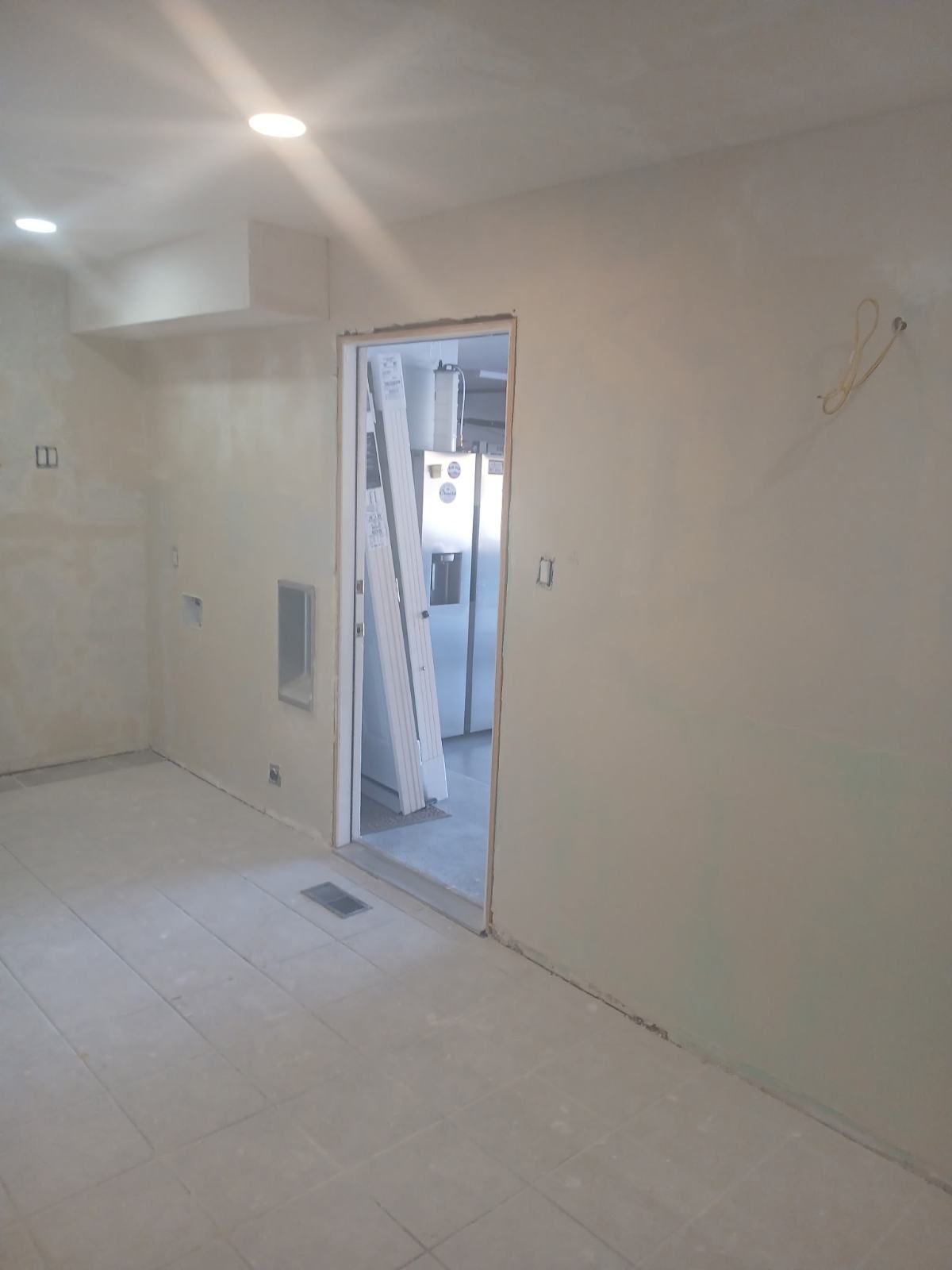 Interior room undergoing renovation; doorway leads to kitchen with refrigerator. Walls are primed, unfinished.