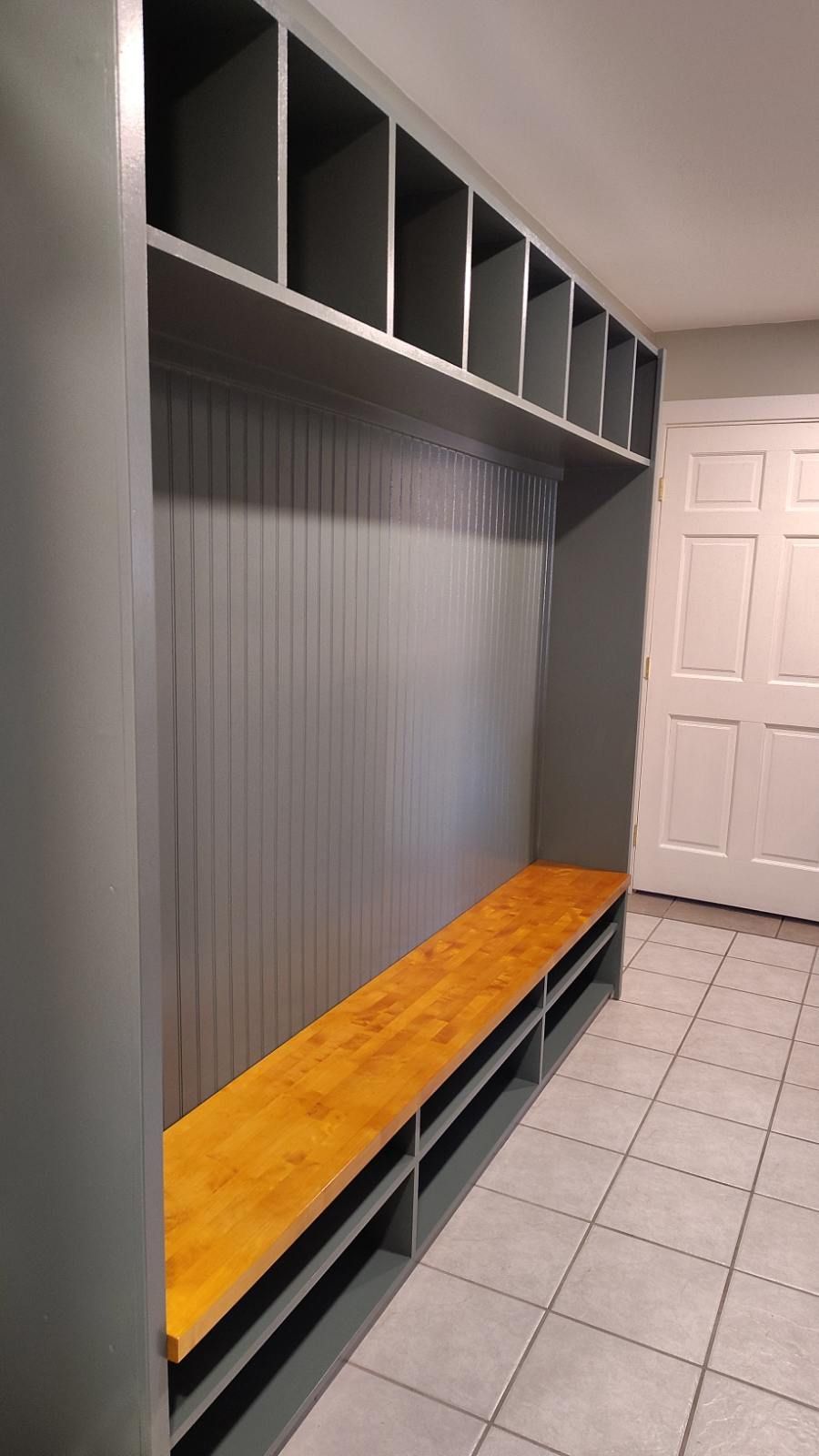 Gray built-in storage unit with cubbies, bench, and lower storage compartments. Wooden seat. White door in background.