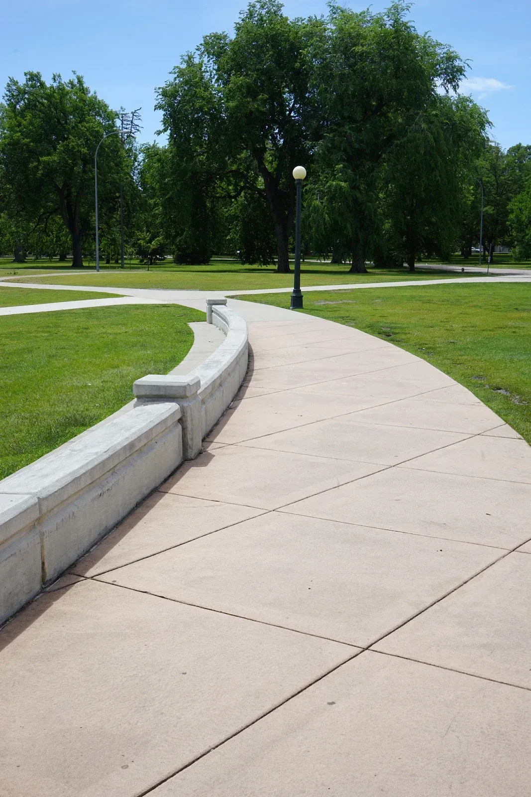 Curving concrete path with low gray wall in a park. Green grass, trees, and a blue sky.