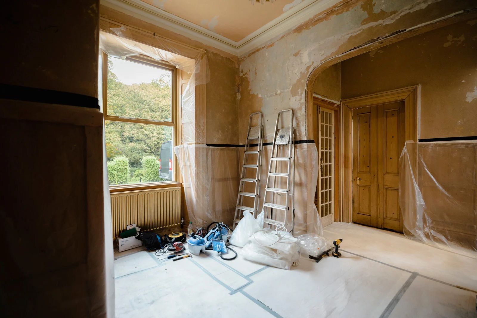 Room under renovation with ladders, tools, and plastic sheeting.
