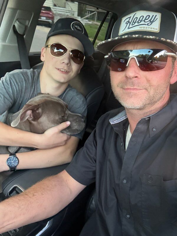 Two people in sunglasses sit in a car, one holding a gray dog; one wears a black cap.