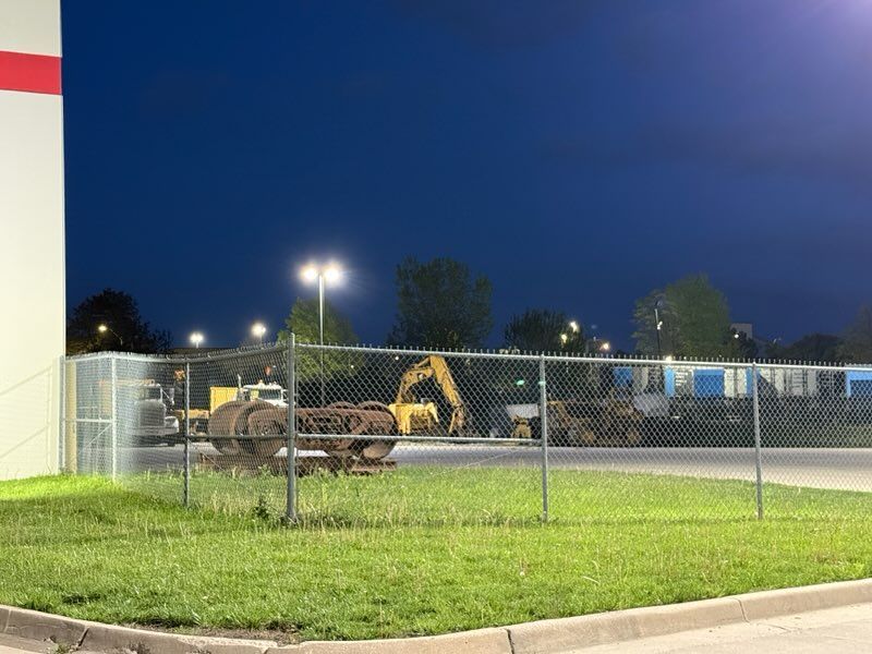 Construction equipment behind a chain-link fence at dusk, with a lit street and grassy foreground.