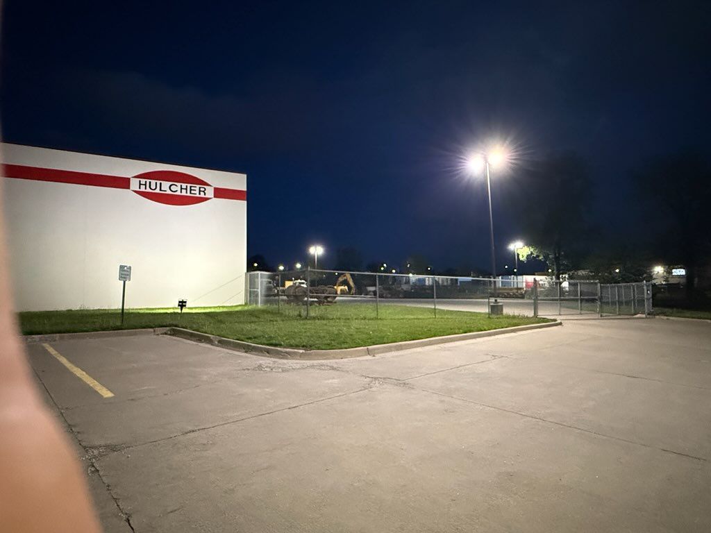 Nighttime view of a lit parking lot beside a white building with a red stripe and sign, near a fenced entrance