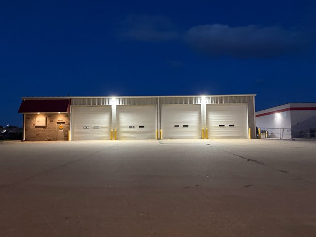 Row of lit garage bays on a building at night, with a large empty paved lot in front.