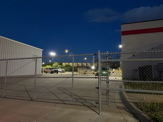 Chain-link gate at a fenced industrial lot at night, with bright lights and parked vehicles.