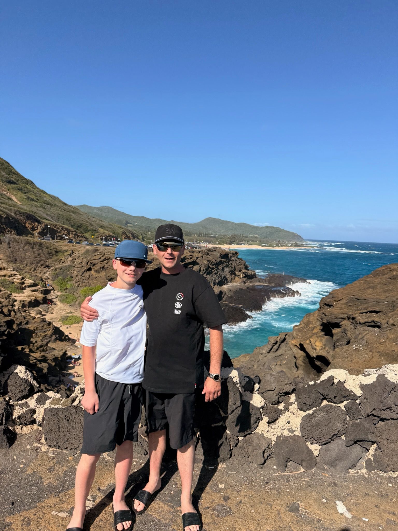 Two people standing on rocky coast with blue ocean and clear sky behind them