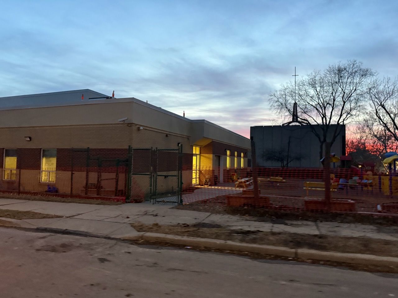 Small building at dusk with lit windows, bare trees, and a fenced patio beside a street.
