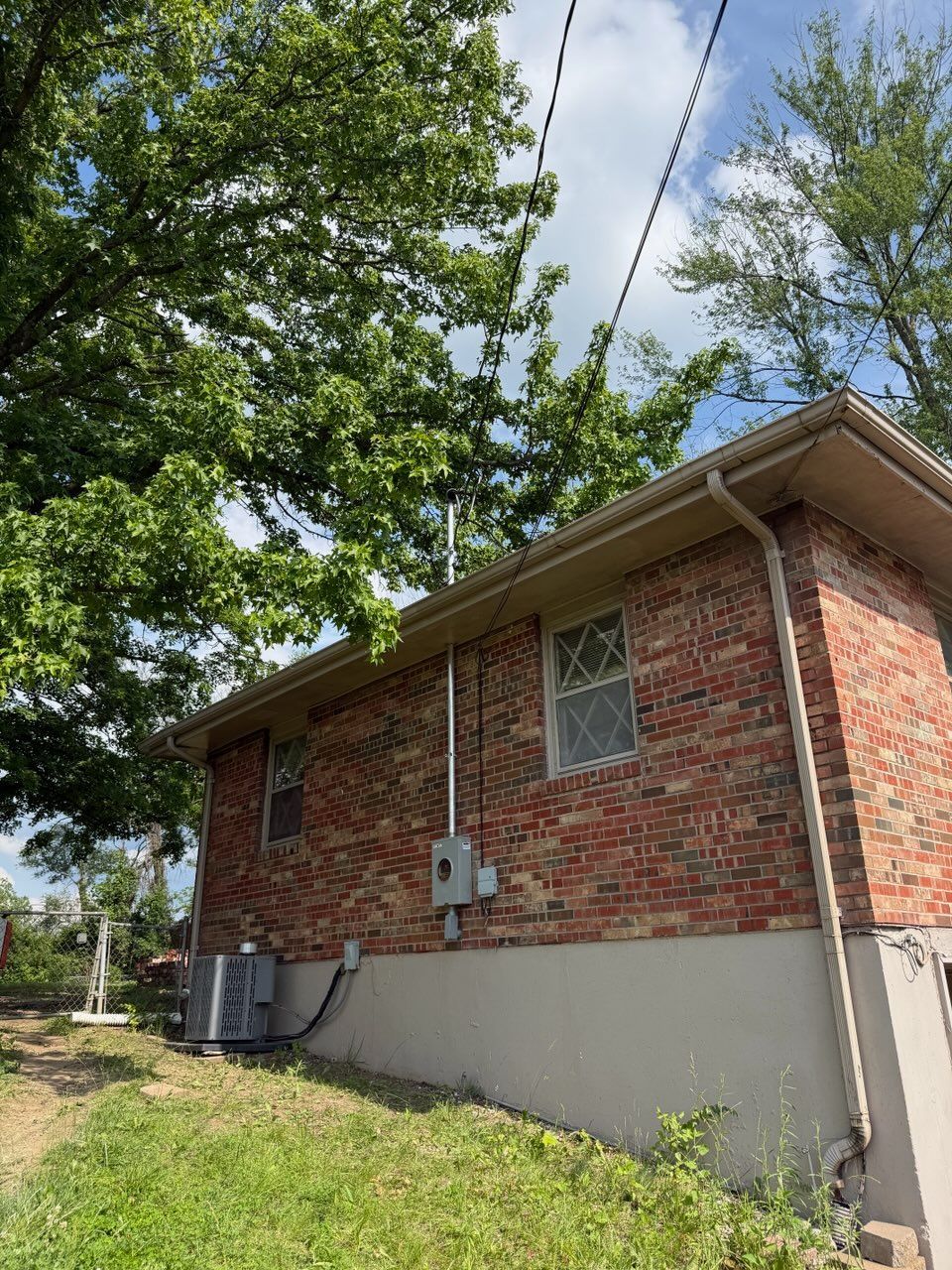 Brick house exterior with gray foundation, utility meters, and trees overhead