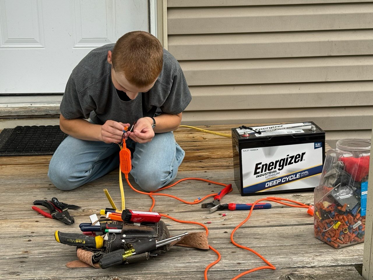 Person kneeling on a driveway, working with wires and tools beside a large battery and assorted cables