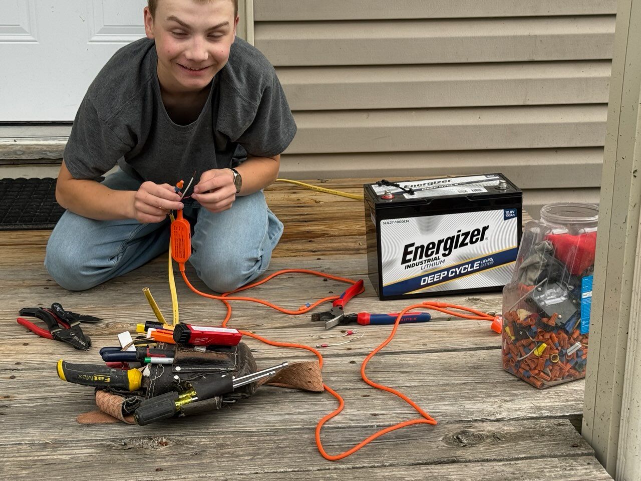 Person kneeling beside a battery setup with orange wires and tools on a porch.