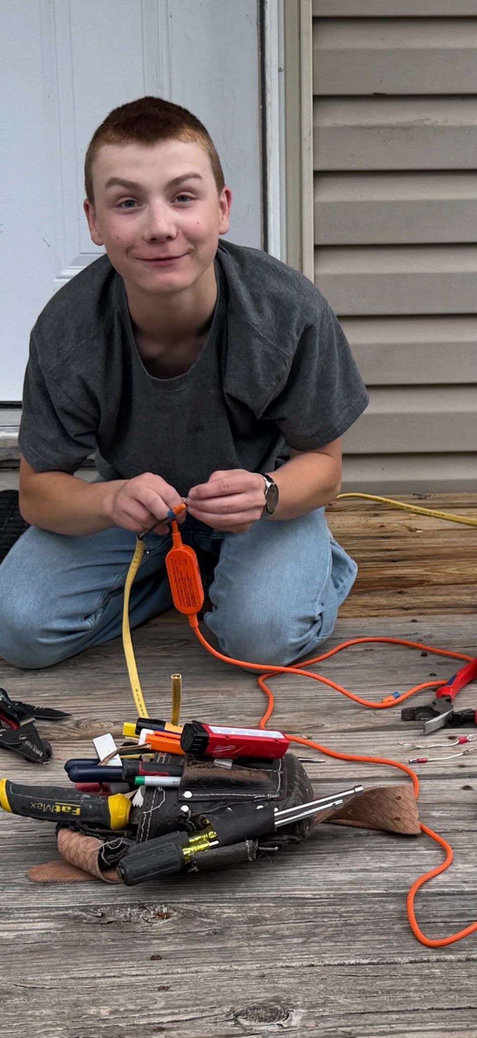 Person kneeling on a wooden deck beside wires and tools, working on a small device with a screwdriver.