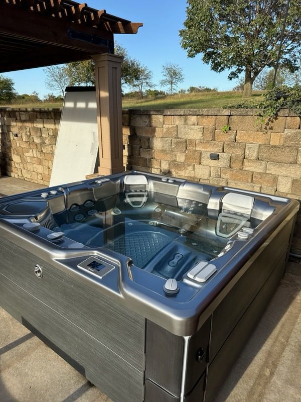 Outdoor hot tub on a patio beside a stone wall, with a pergola and trees in the background.