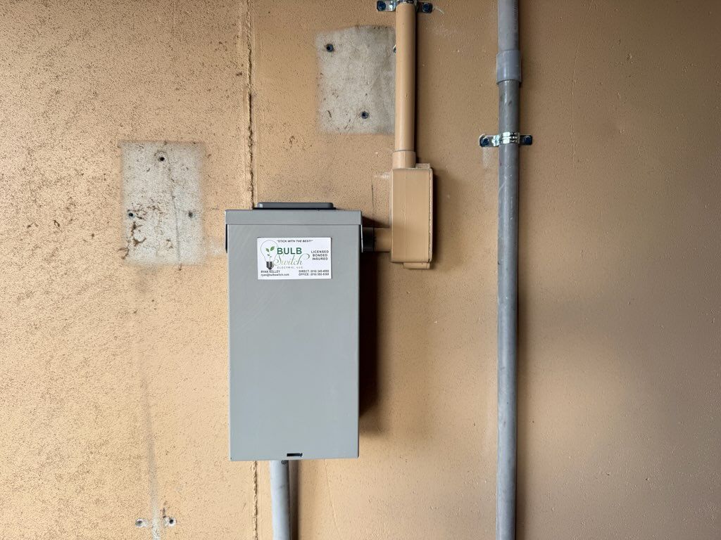 Gray electrical box mounted on a beige exterior wall beside a vertical metal pipe