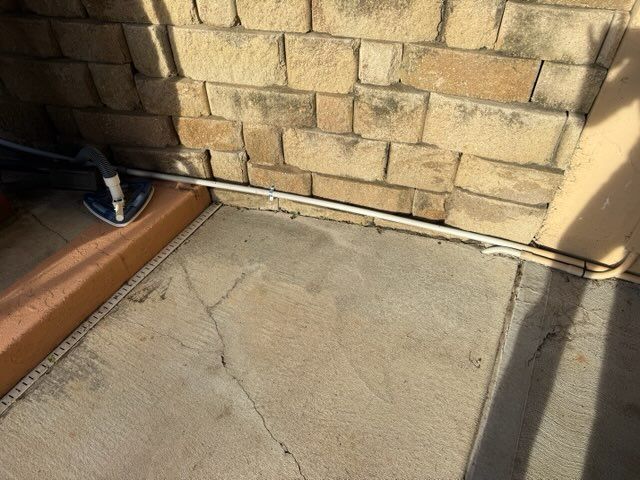 Cracked concrete patio beside a brick wall with a person’s shoe at the edge and strong shadows.