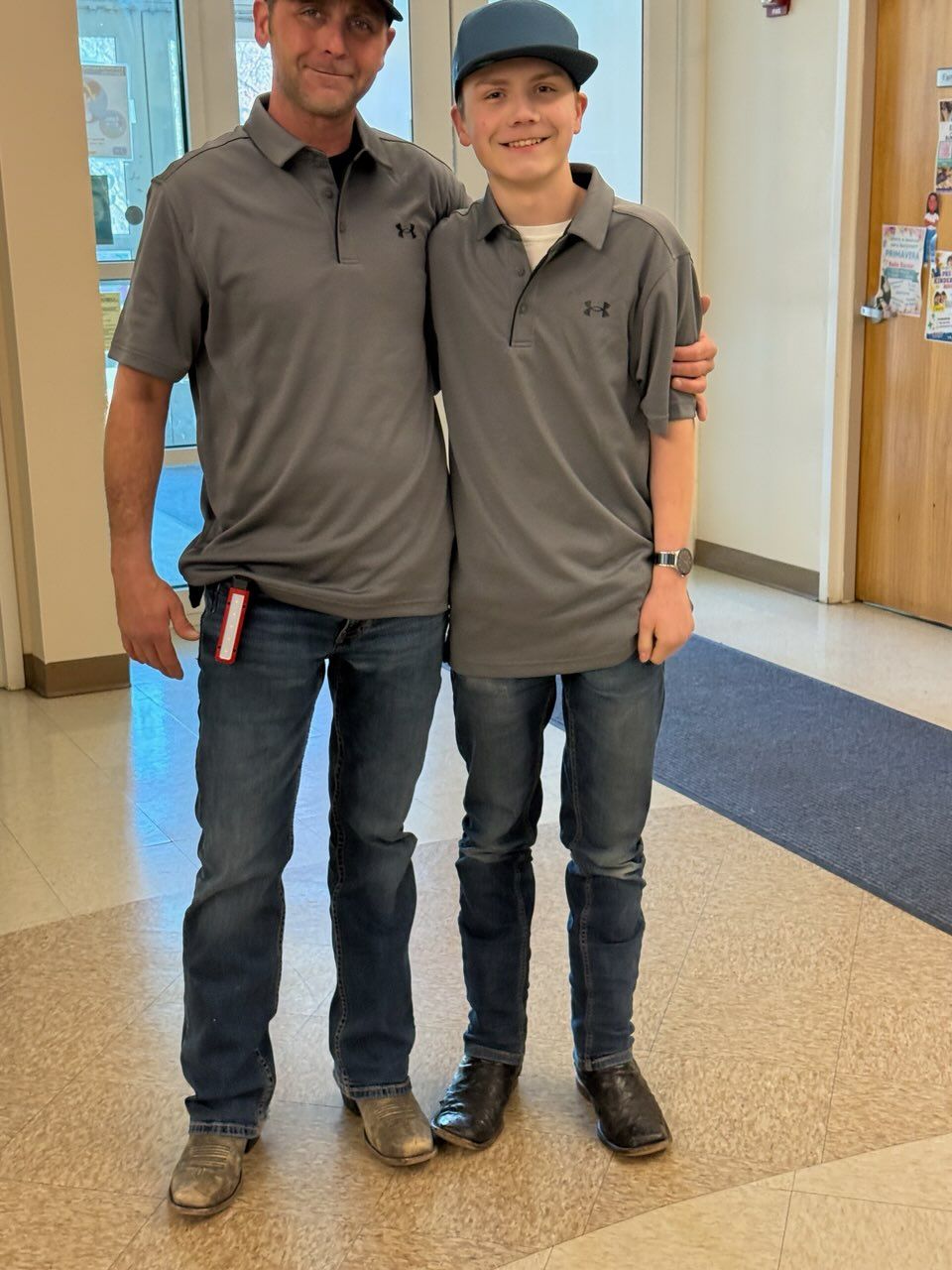 Two people standing in a hallway, wearing matching gray shirts, jeans, and baseball caps.