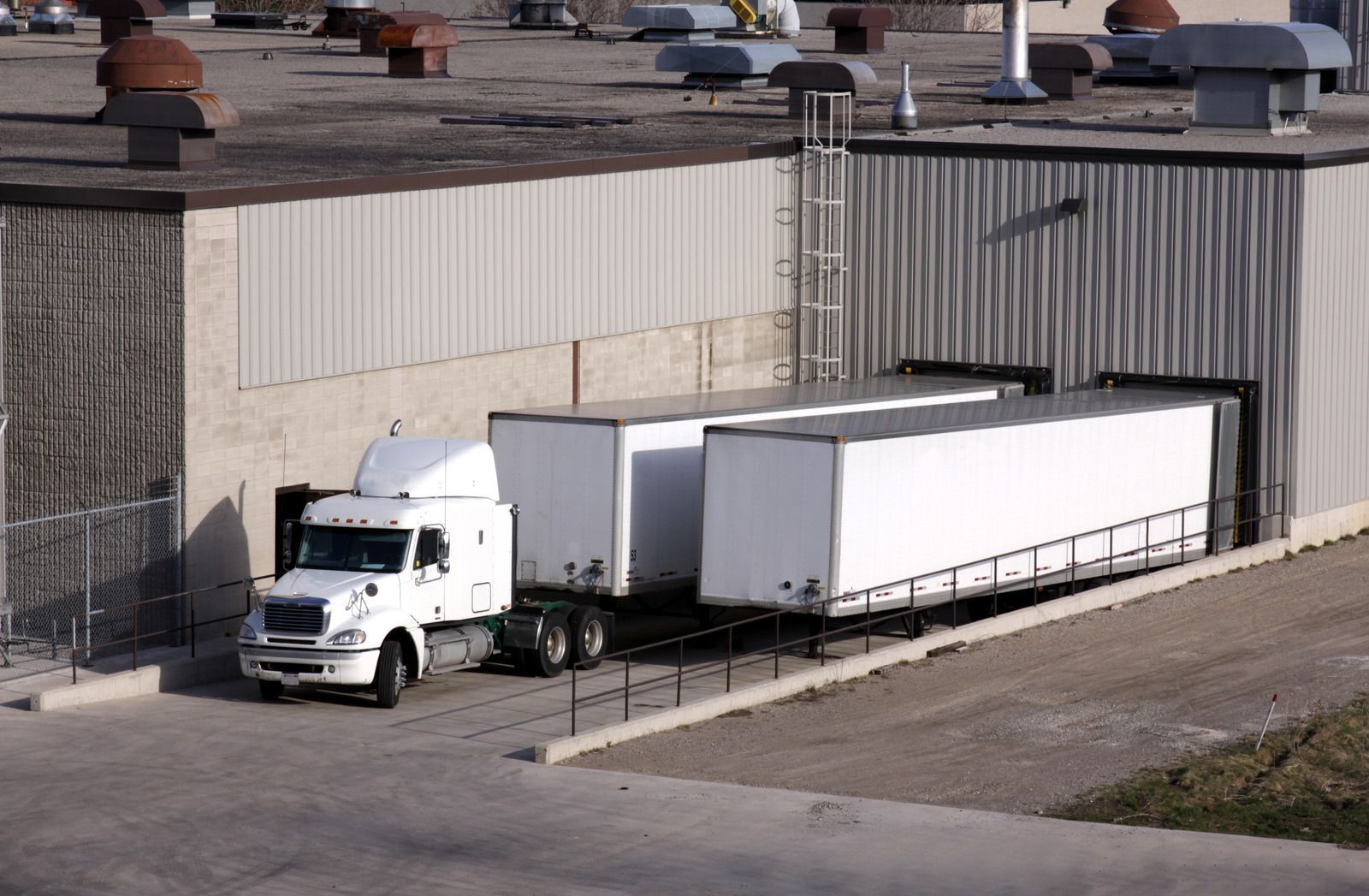 A white semi-truck is at a loading dock with two attached white trailers.