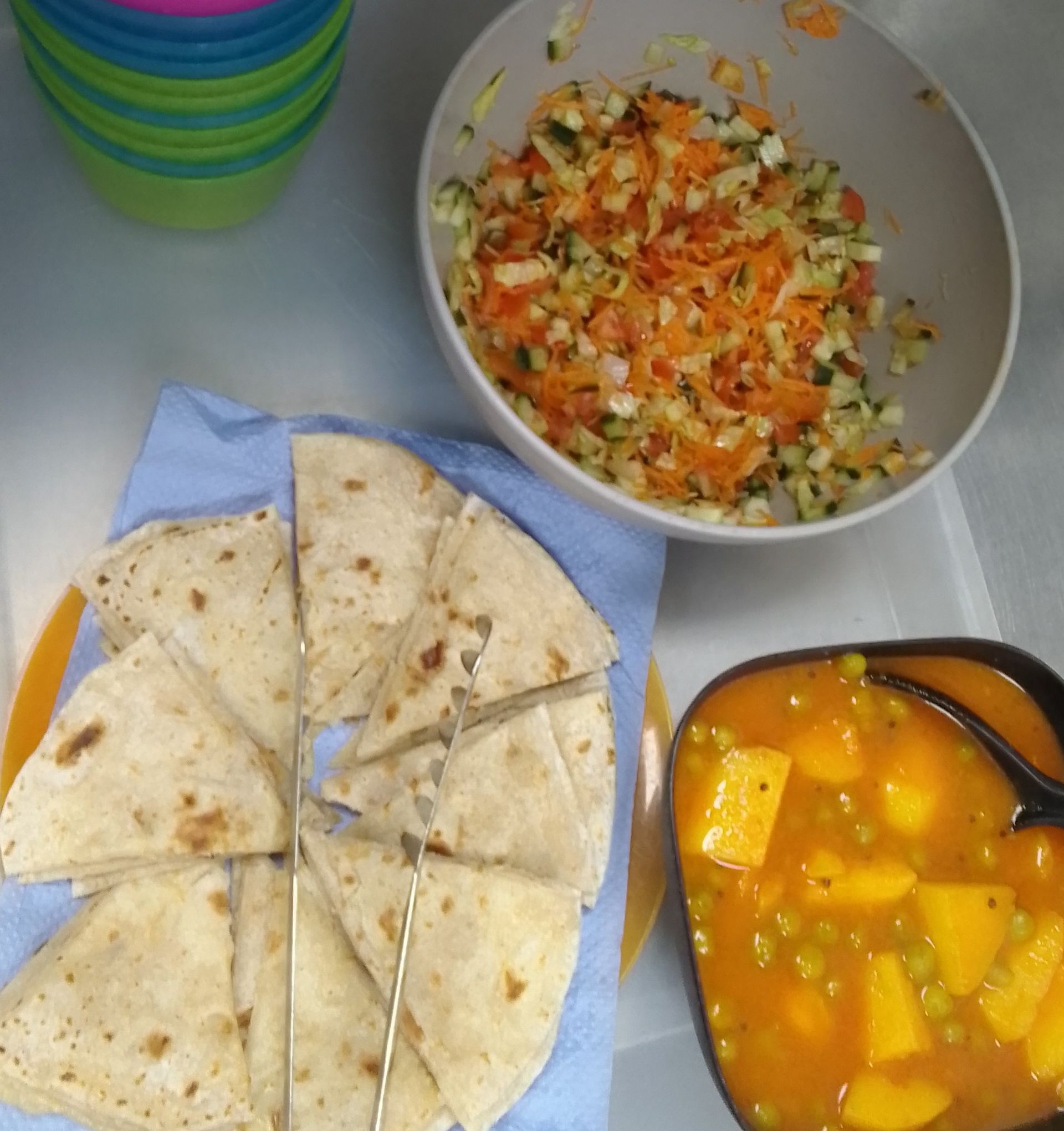 Flatbread wedges, chopped salad, and a bowl of orange curry on a tray with stacked cups.