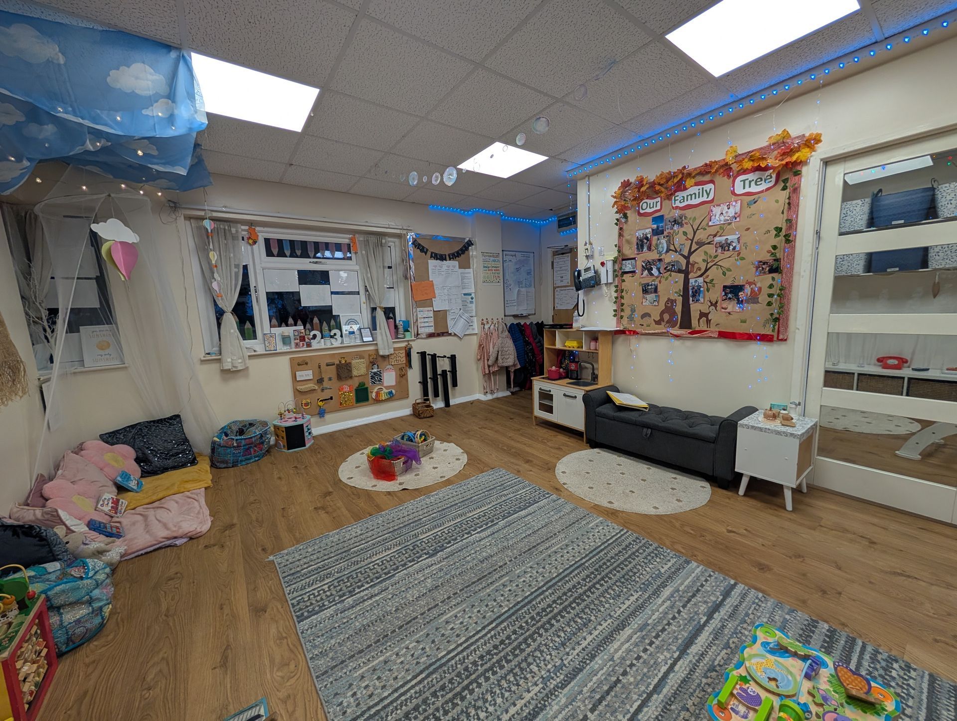 Colorful classroom with rugs, shelves, and a bulletin board, lit by bright ceiling lights.