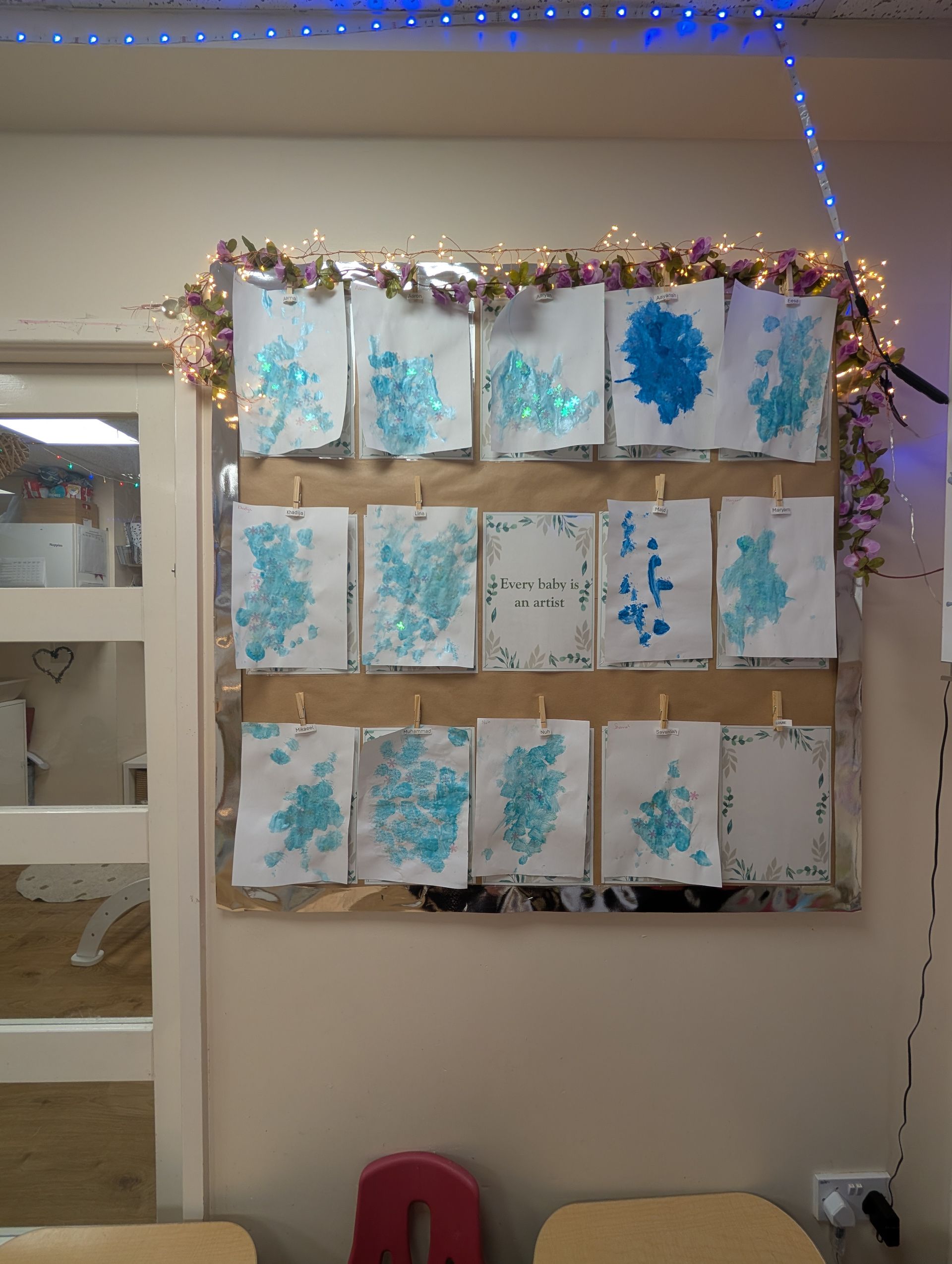 Wall display of blue watercolor paintings framed with holiday garland and string lights in a classroom.
