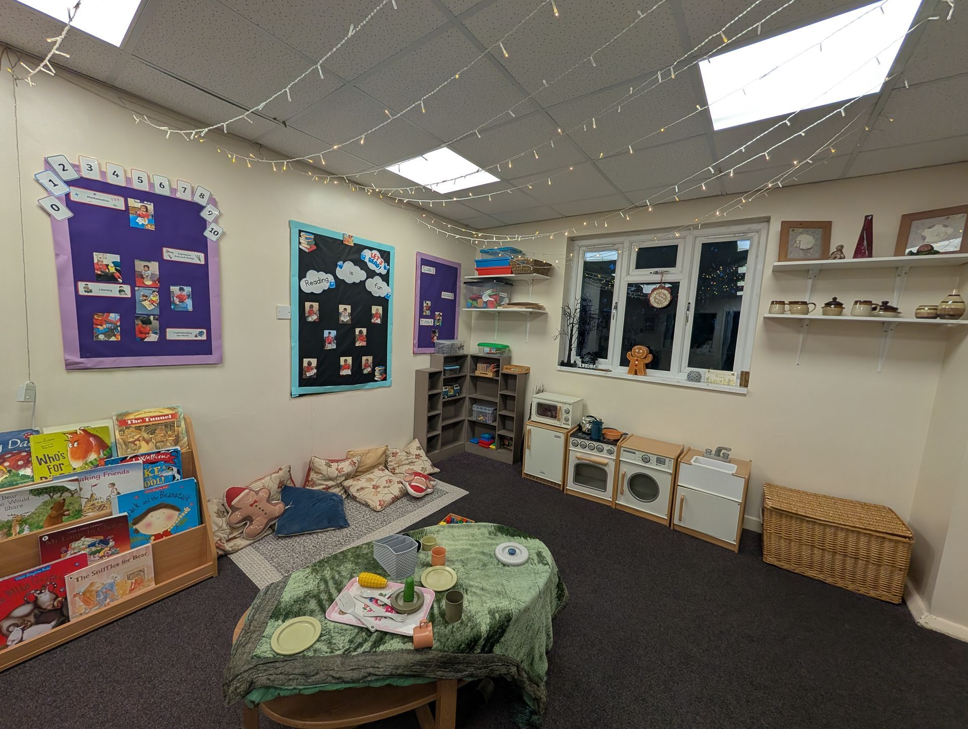 Small classroom reading corner with low green table, shelves, wall posters, windows, and woven baskets.