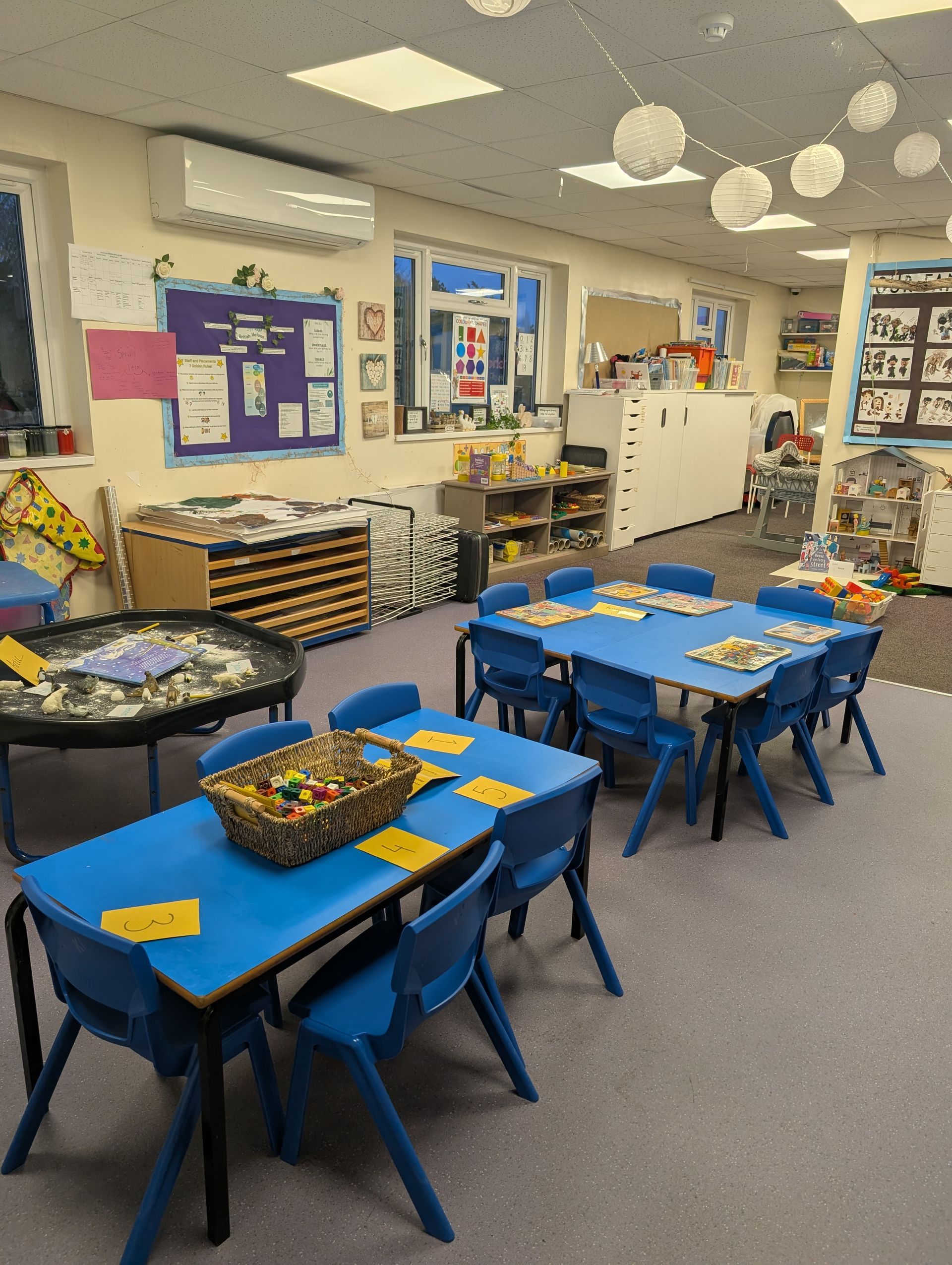 Classroom with blue tables and chairs, baskets of materials, and bulletin boards along the walls.