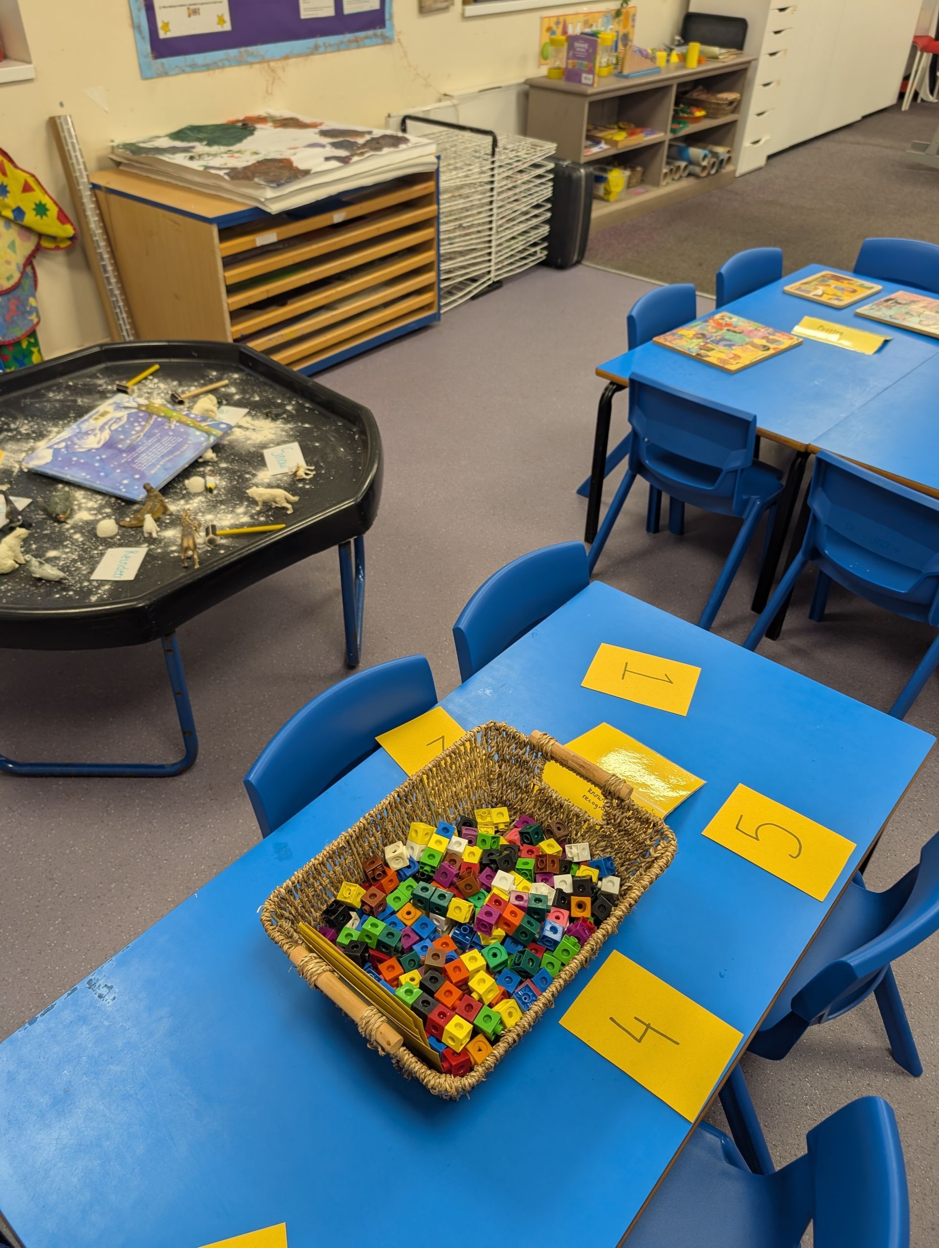 Classroom with blue tables and chairs, baskets of colorful blocks, and shelves along the wall