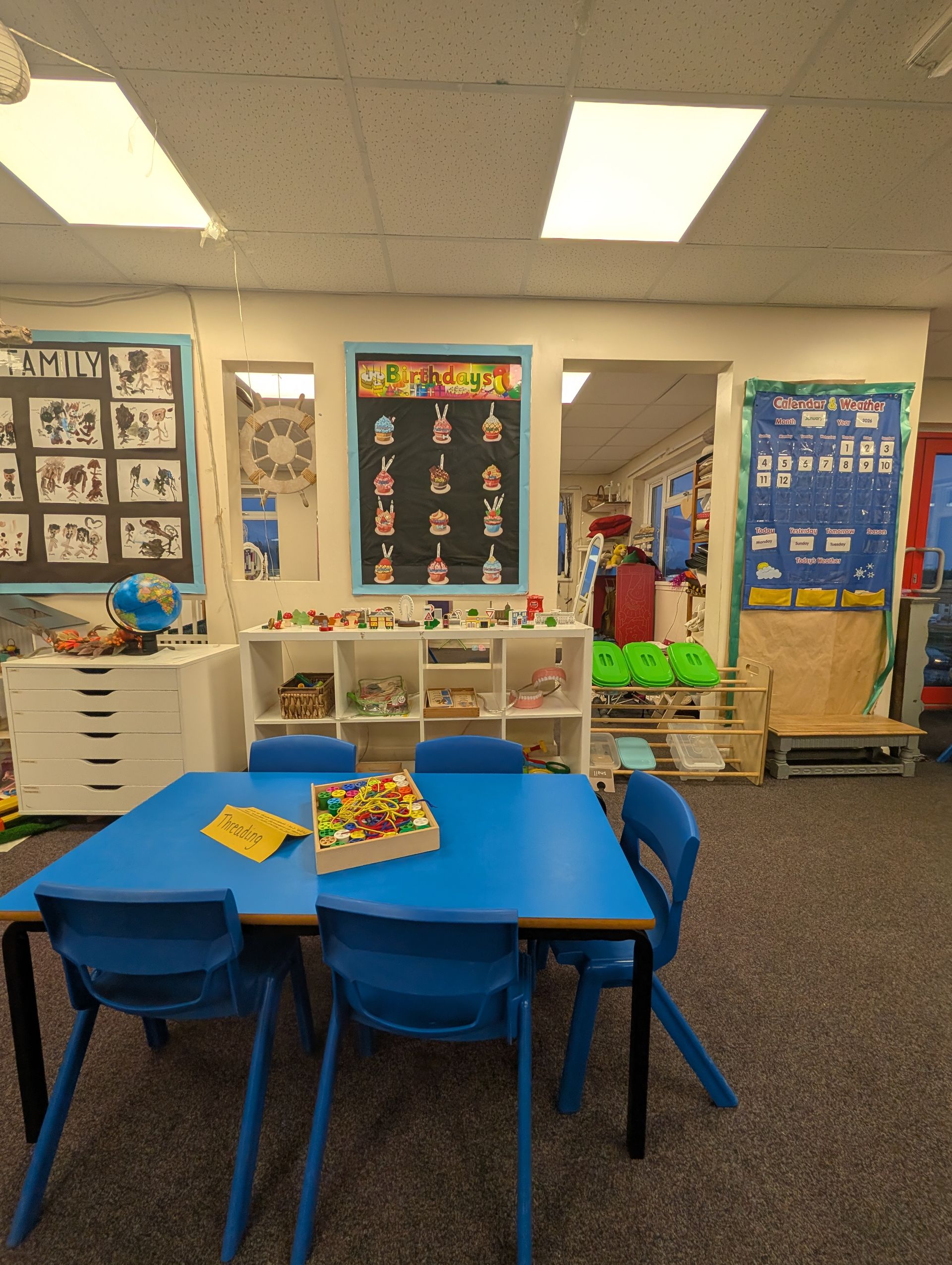 Classroom with blue tables and chairs, bulletin boards, and shelves of books and supplies under bright ceiling lights