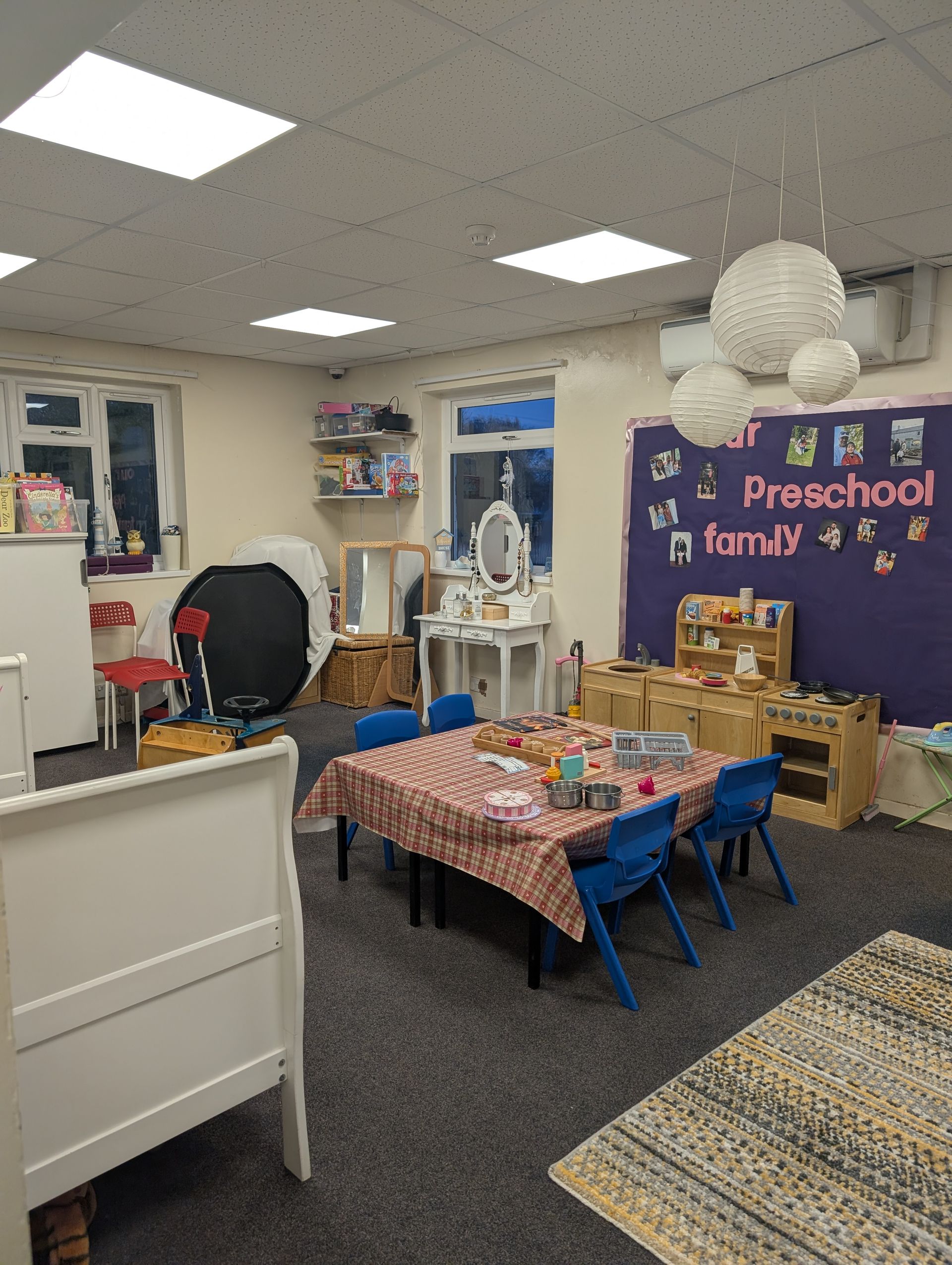 Classroom reading nook with small table, blue chairs, bookshelves, and a “Pre-school” bulletin board