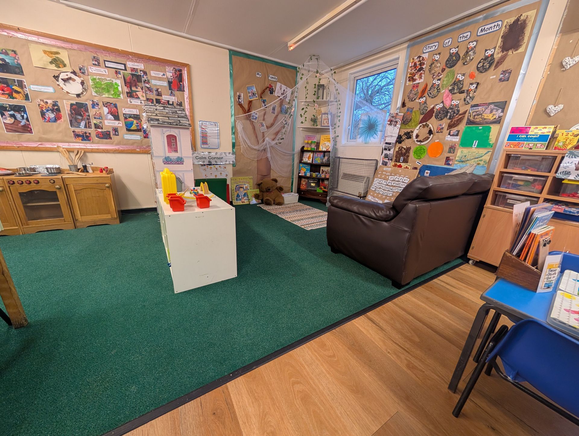 Colorful classroom with carpeted play area, bulletin boards, a brown chair, and shelves of books and supplies.