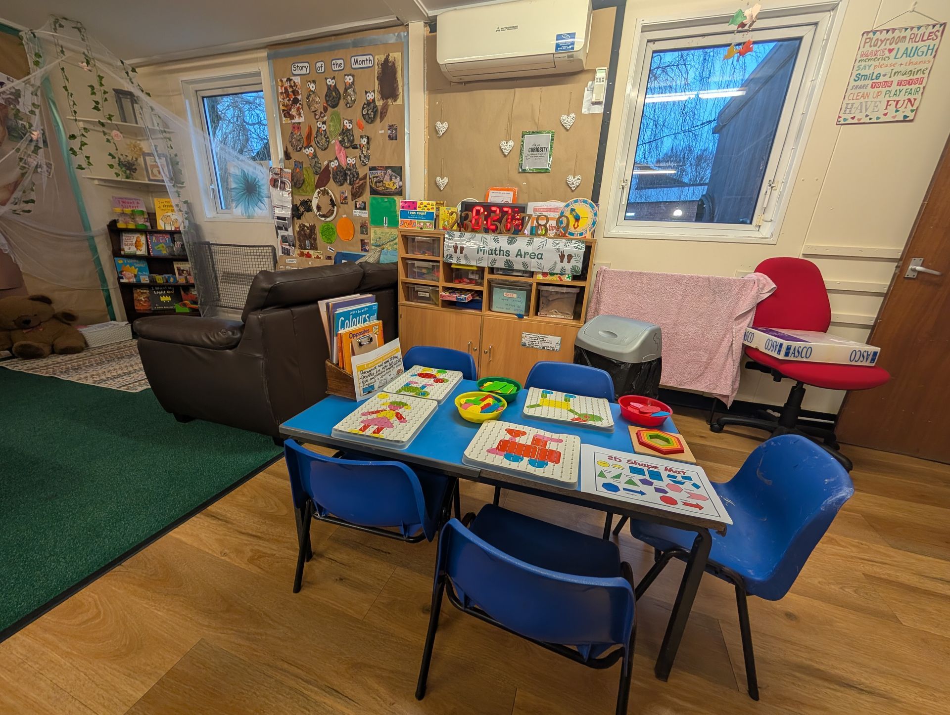 Small classroom with children’s tables, blue chairs, toy shelves, and a wall-mounted air conditioner