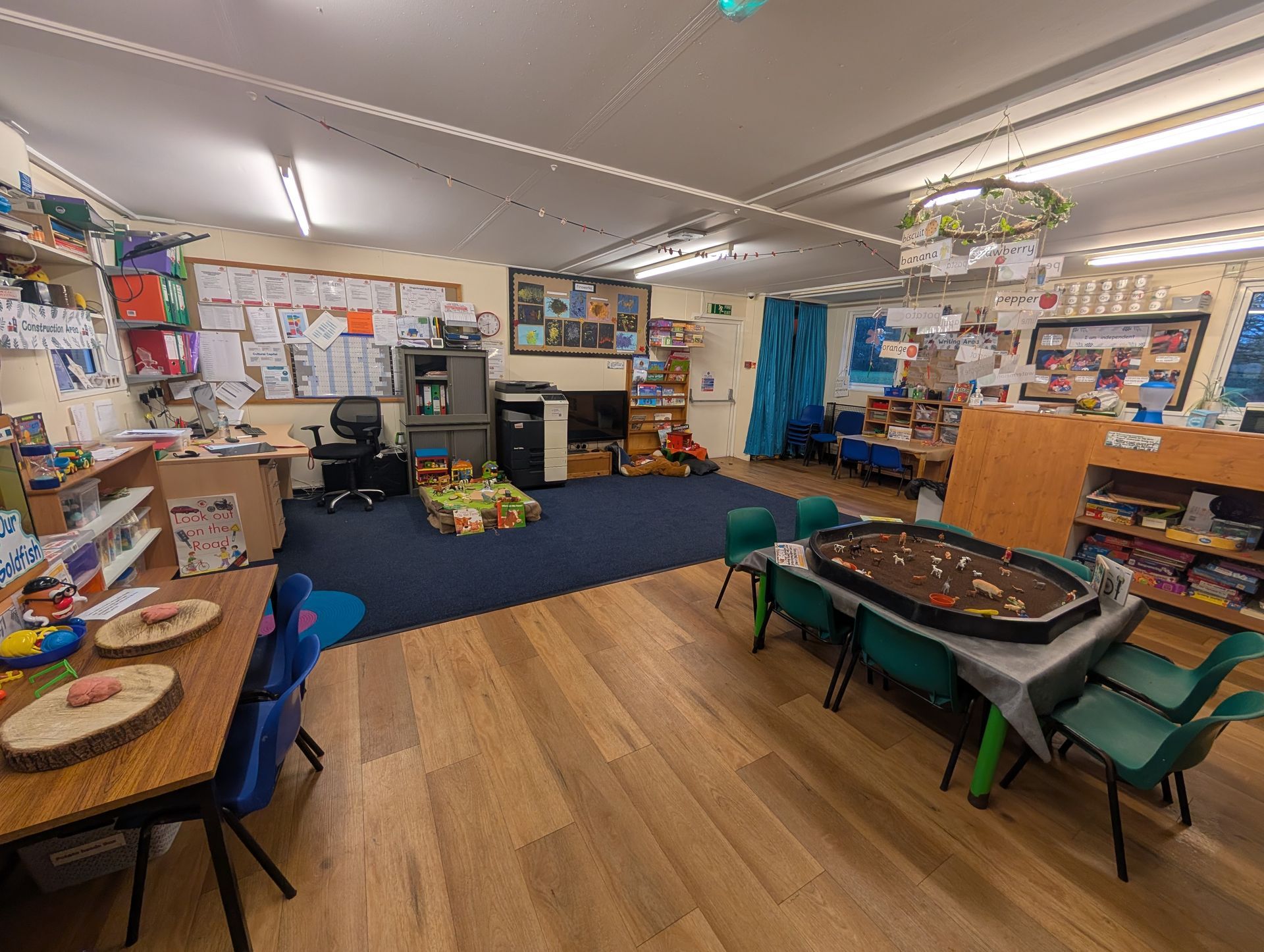 Classroom with tables, chairs, artwork, bookshelves, and a blue carpeted reading area