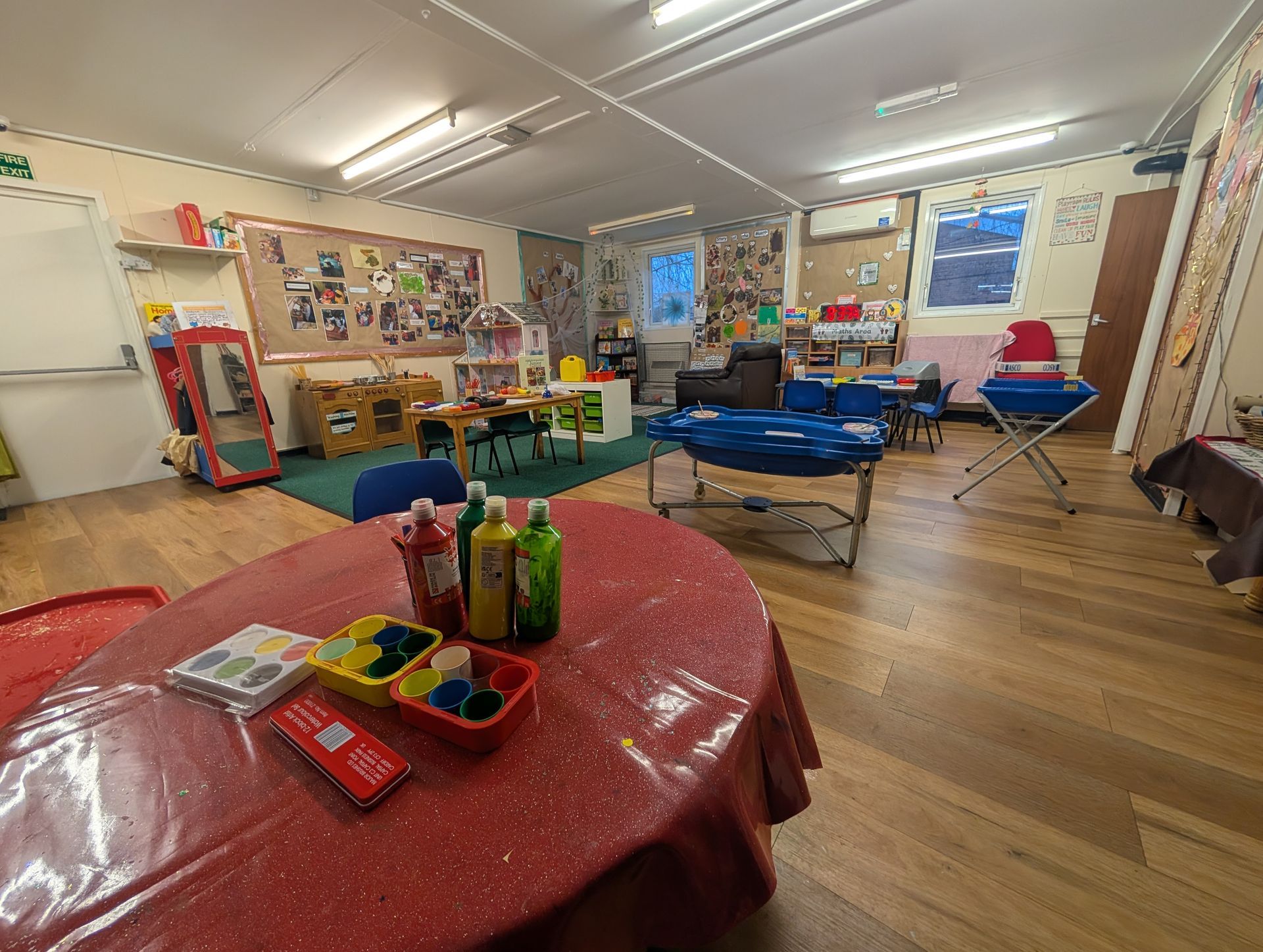 Classroom with tables, chairs, toys, and colorful posters, lit by overhead lights