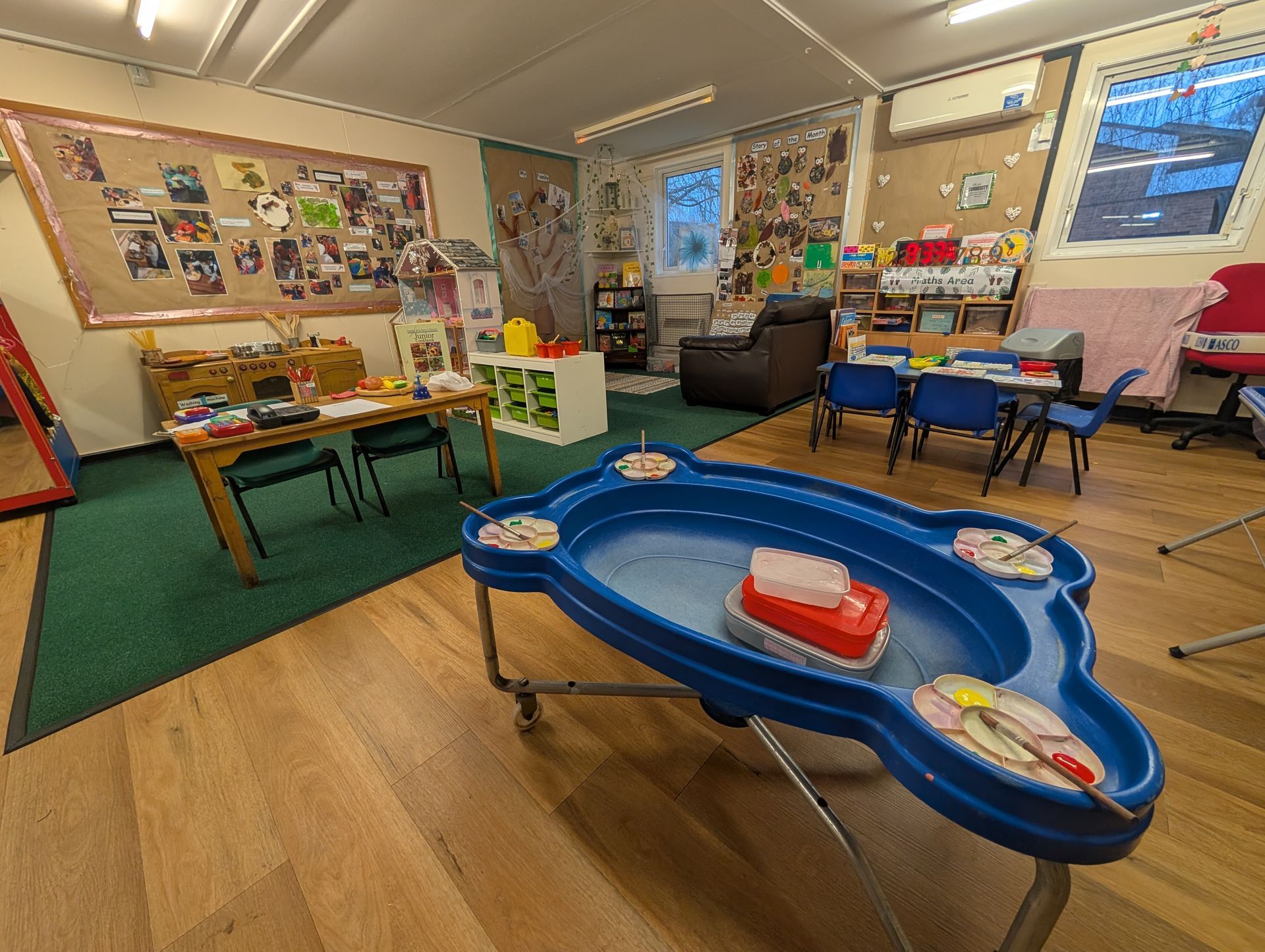 Bright classroom with toys, tables, chairs, and learning materials spread across the room.